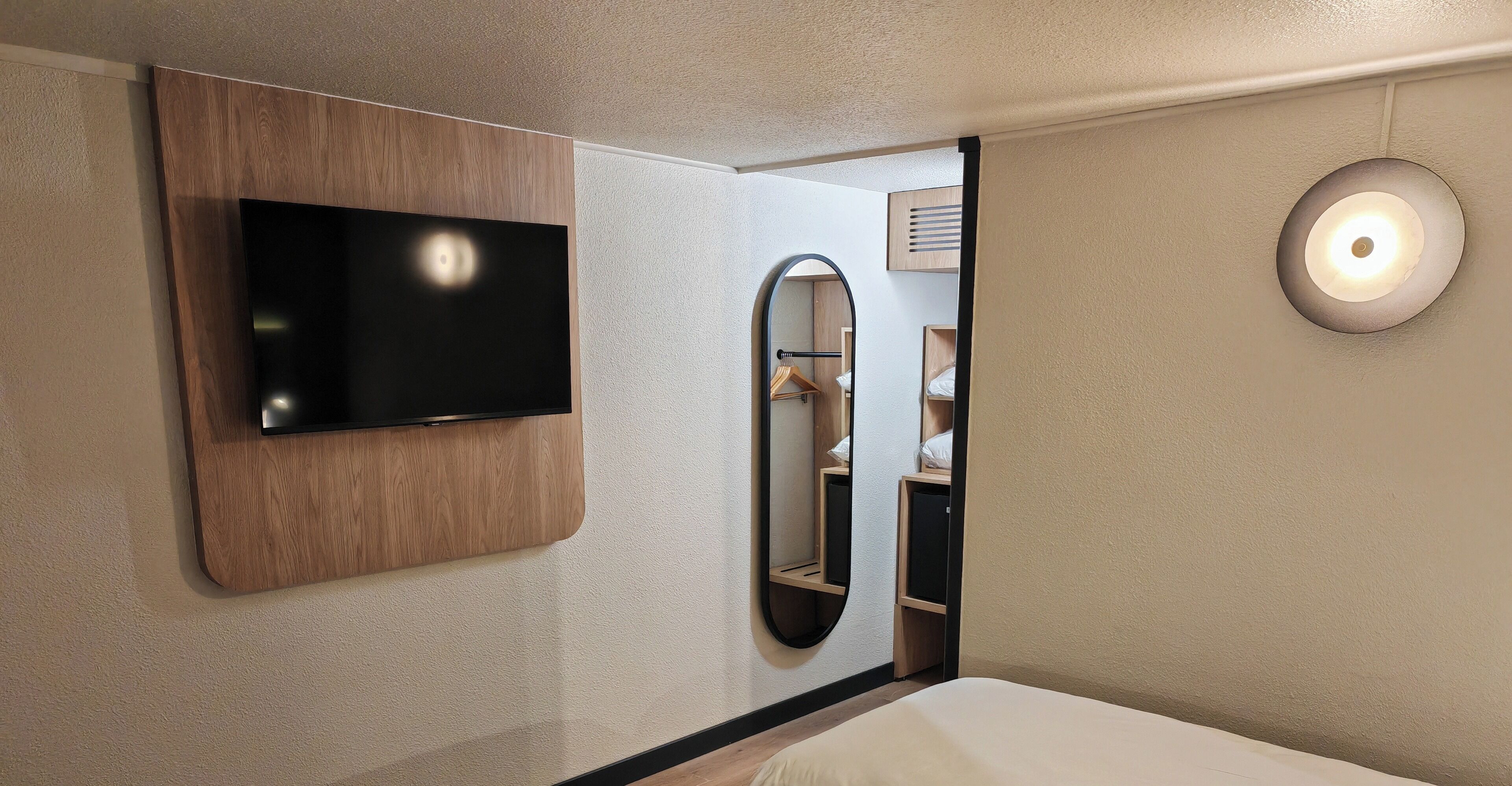 A hotel room with a mounted TV, a mirror, and a bedside lamp.