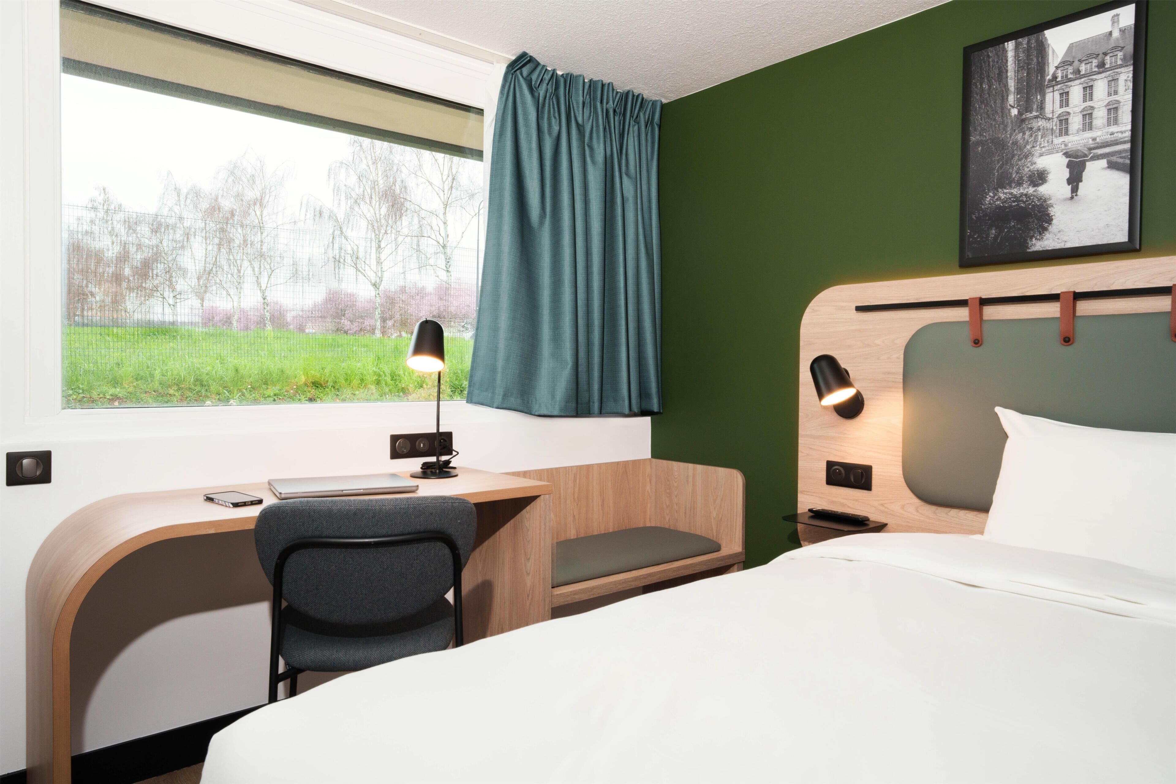 Een hotelkamer met een bed, bureau, stoel en uitzicht op een groen landschap door een raam.