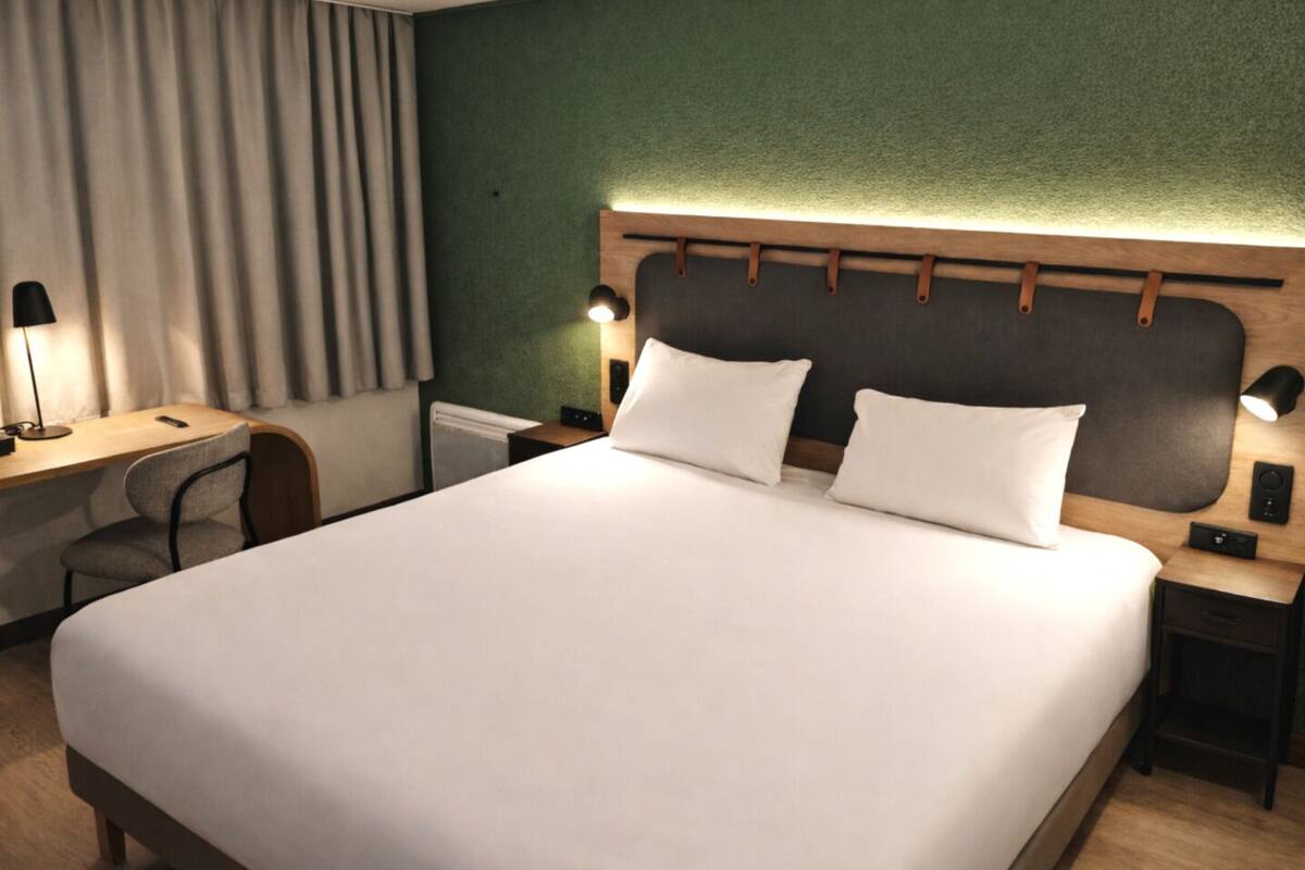 A neatly arranged hotel room with a large bed, a desk with a chair, a nightstand with a lamp, and a headboard with a light fixture.