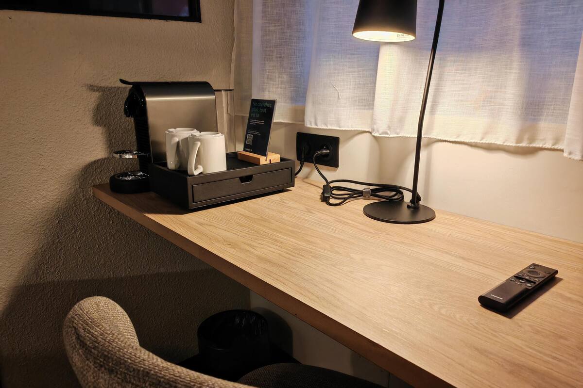 A wooden desk with a coffee machine, a remote control, and a lamp.