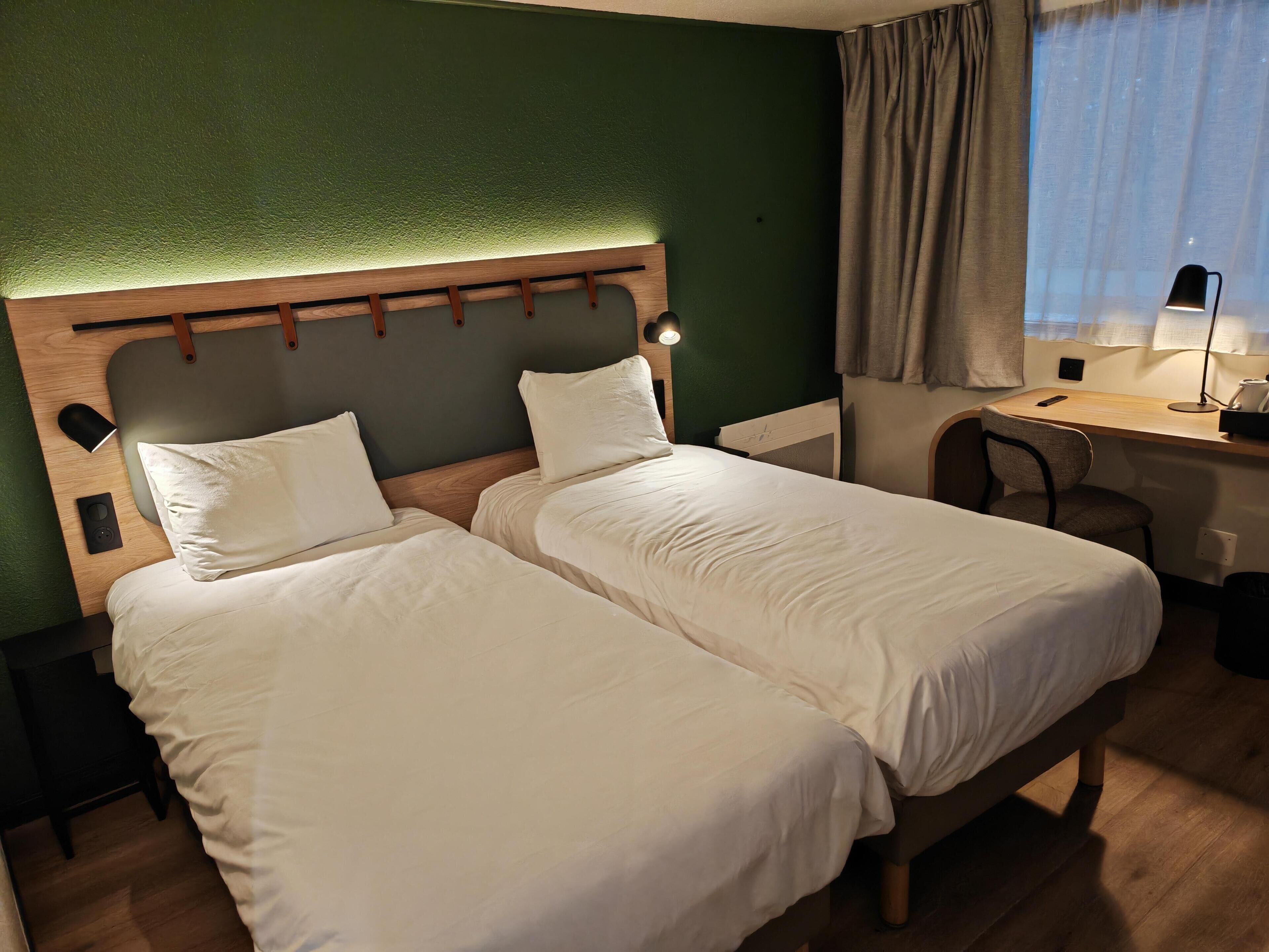 A hotel room with two beds, a desk, a chair, and a window with curtains.