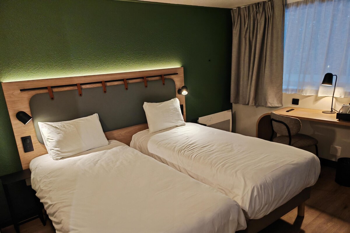 Une chambre d’hôtel avec deux lits, un bureau, une chaise et une fenêtre avec des rideaux.