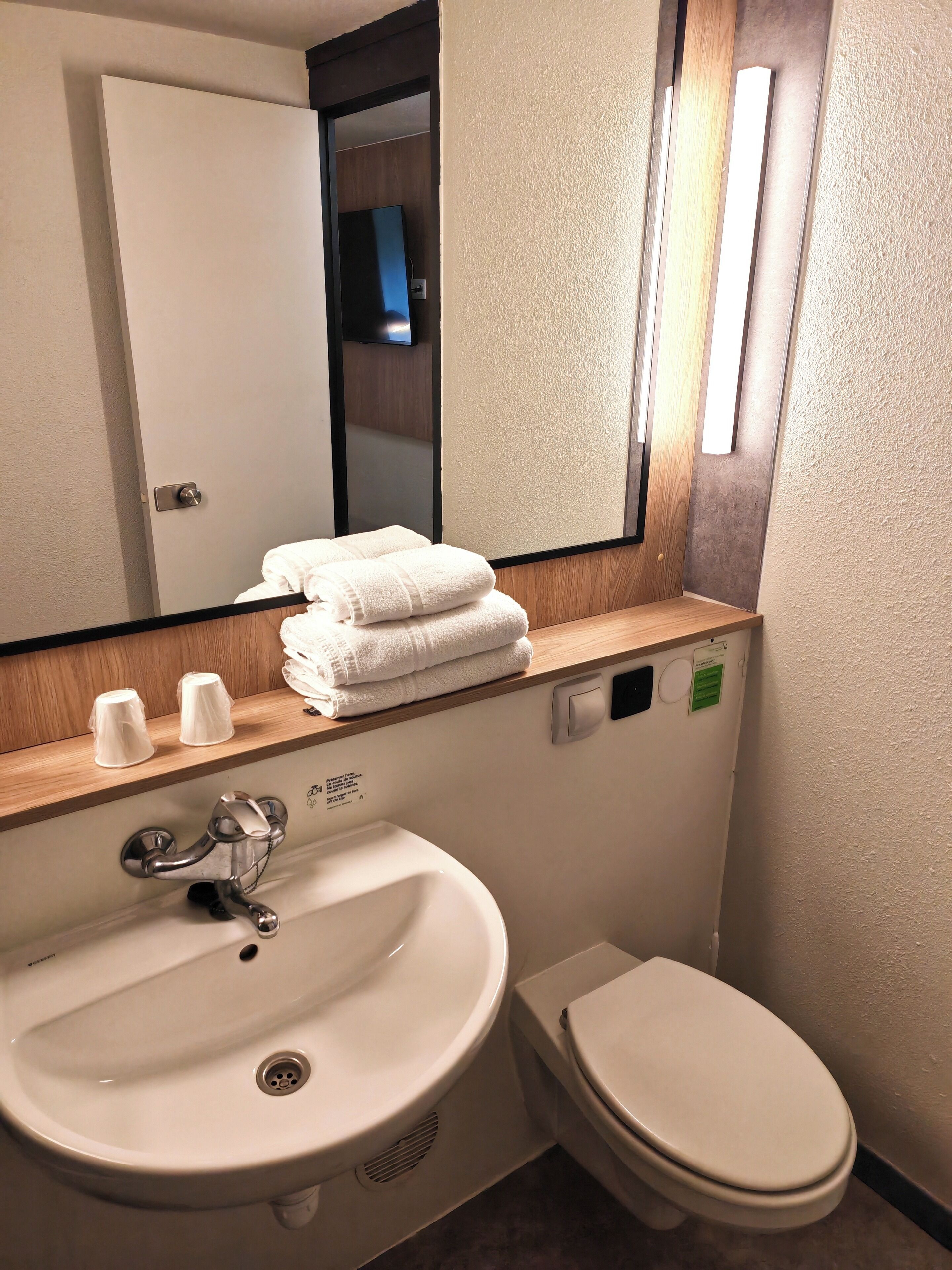 Une salle de bain avec un lavabo, des toilettes et un miroir au-dessus du lavabo. Des serviettes sont pliées et posées sur le bord du lavabo, et une petite étagère contient des tasses.