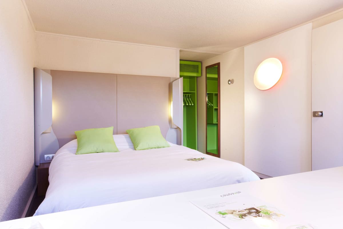 A hotel room with a bed, green pillows, a nightstand, a bedside table, a wall-mounted lamp, a green-framed mirror, and a door with a light fixture.