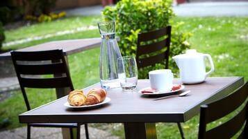 Une table avec un croissant, une carafe, une tasse et un verre d’eau, disposée à l’extérieur, avec des chaises et de la verdure en arrière-plan.