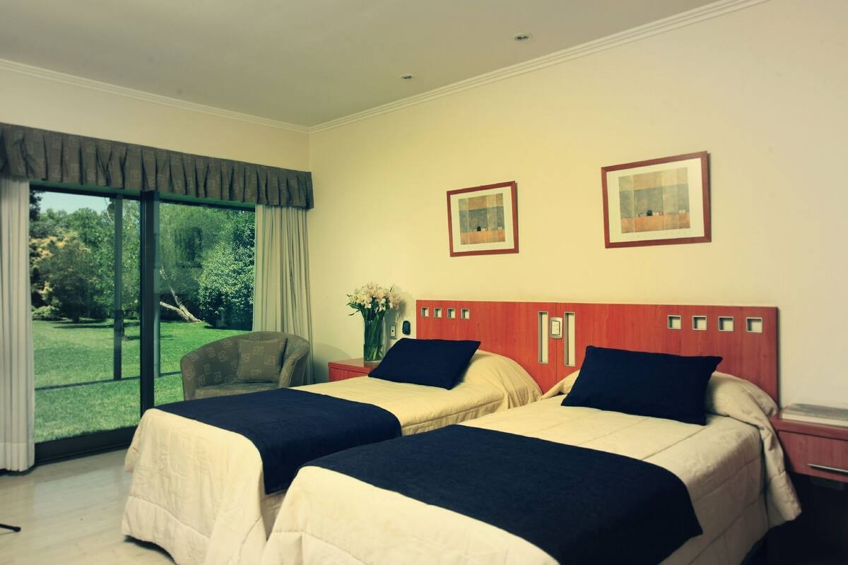 A hotel room with two beds, a large window with a view of greenery, and two framed pictures on the wall.
