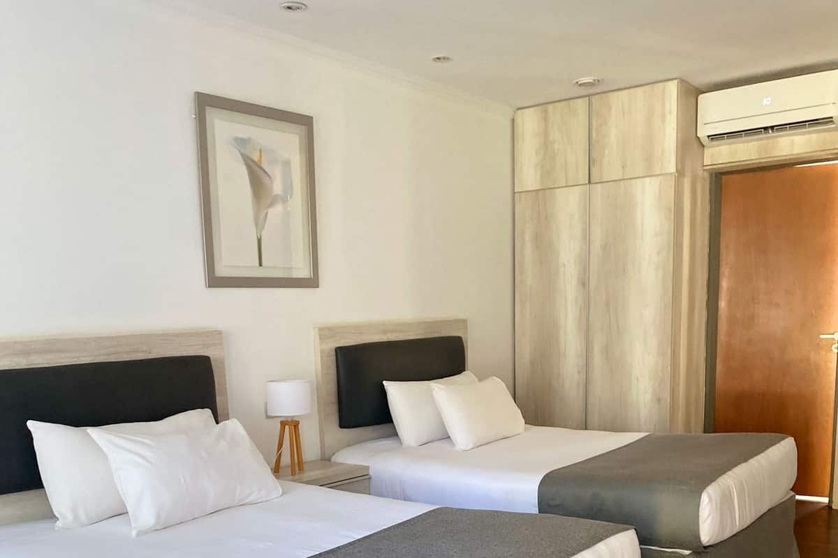 A hotel room with two beds, a nightstand, a lamp, a framed picture on the wall, and an air conditioning unit.
