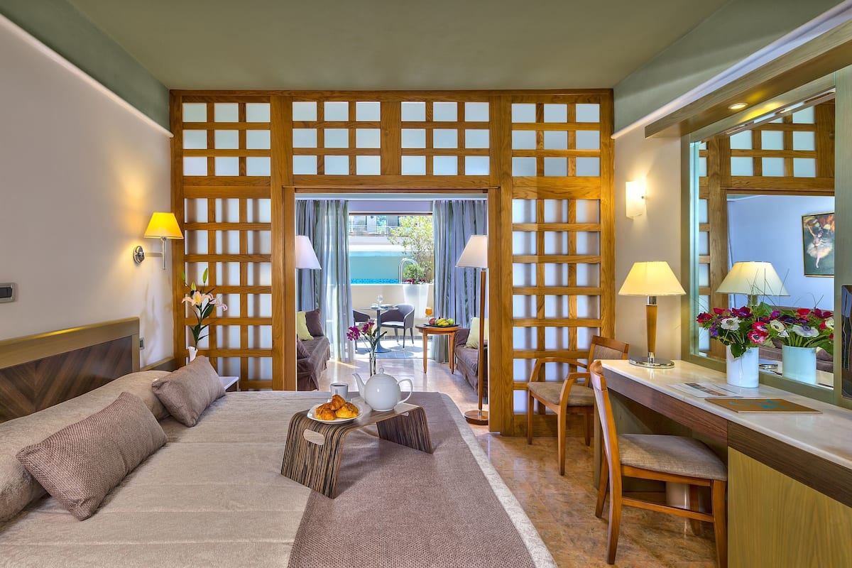 Ein Hotelzimmer mit Bett, Schreibtisch, Stuhl, einem kleinen Tisch mit Teekanne und Speisen sowie Blick auf den Pool und die Außenbereichs-Sitzgelegenheiten durch eine Holzwand.