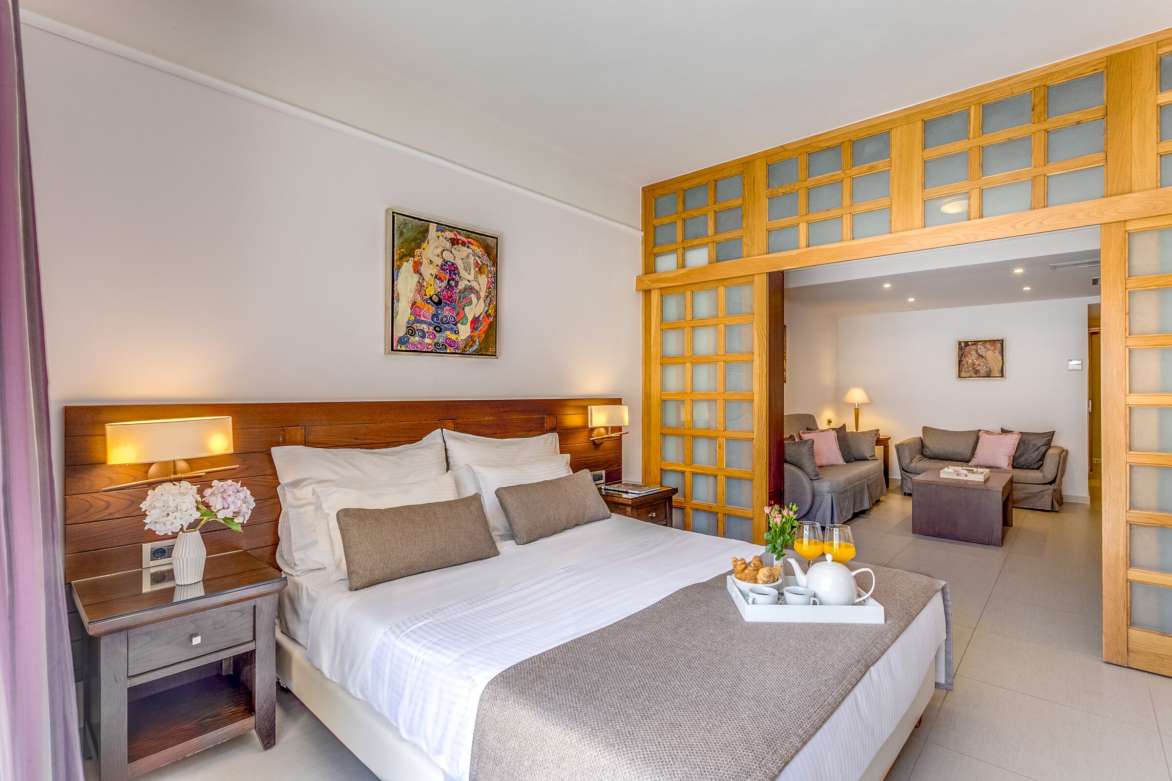 Habitación de hotel con una cama grande, una mesita de noche con lámpara, un cabecero de madera, un cuadro en la pared y una puerta corrediza que da a una sala de estar con sofá y mesa de centro.