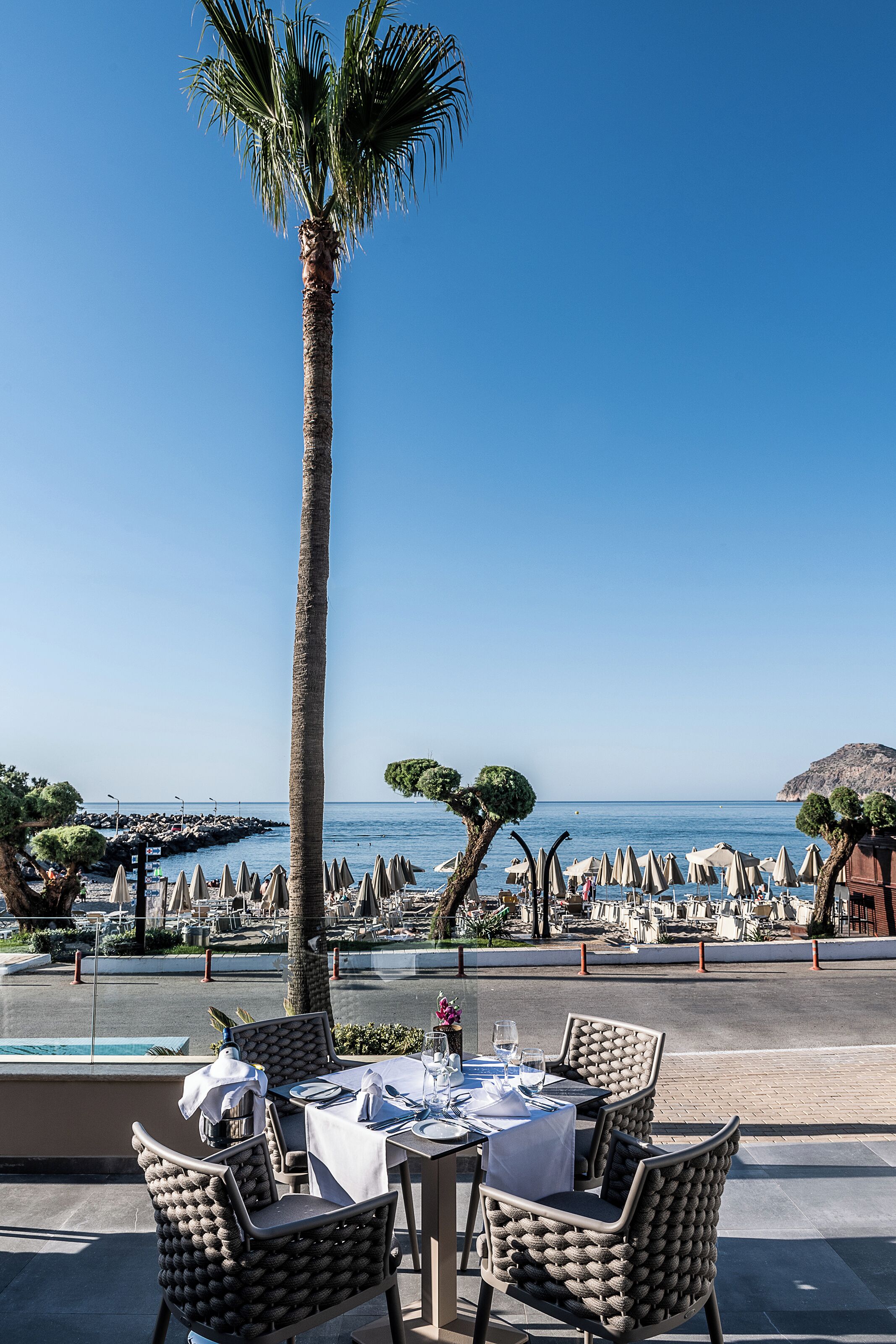 An outdoor dining area with wicker chairs and tables set up for guests, overlooking a beach and the sea.