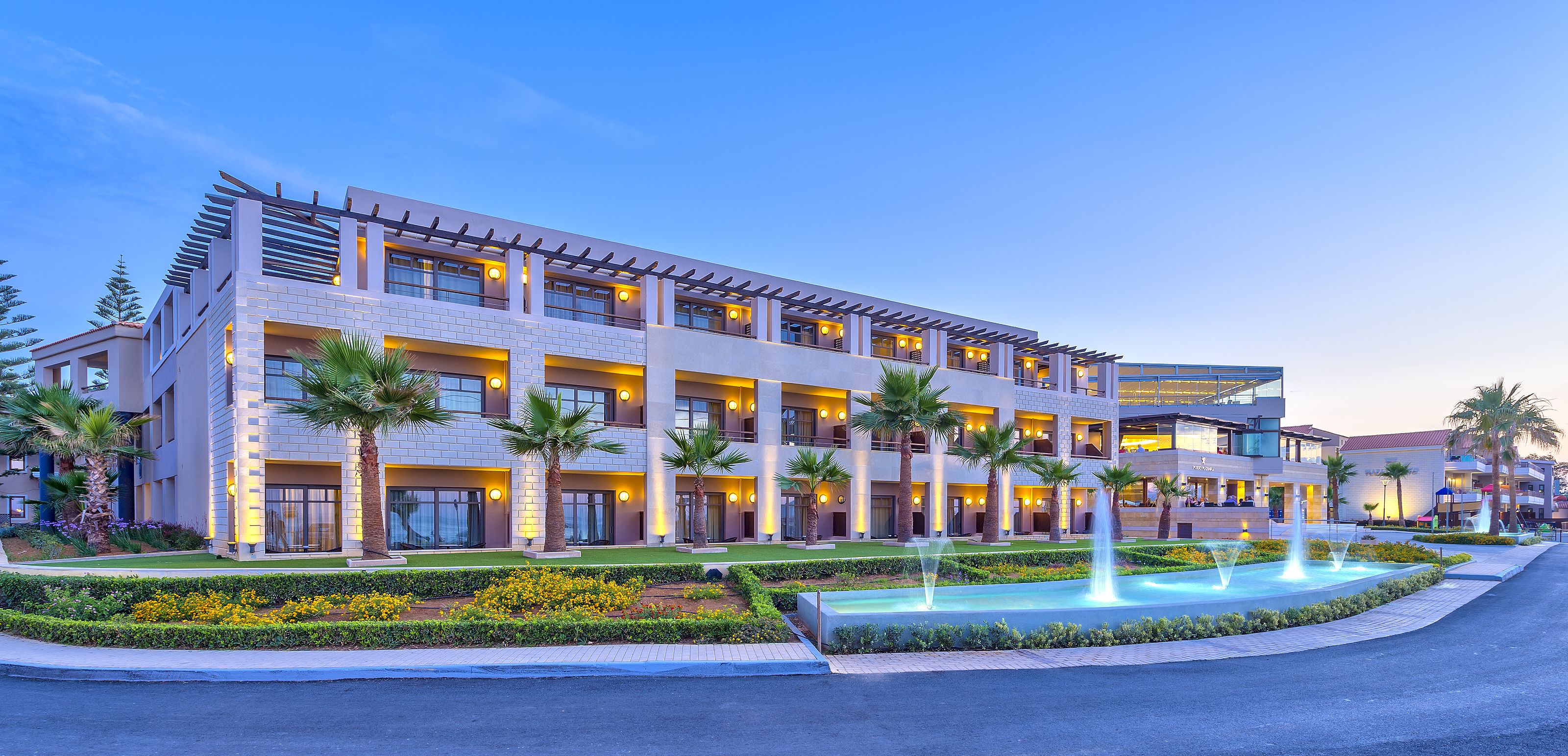 A modern hotel with multiple floors, outdoor lighting, palm trees, and a water feature.