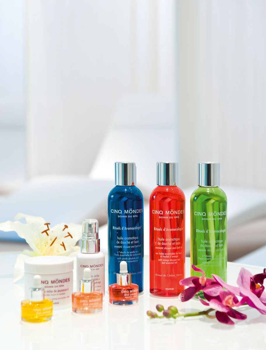 A selection of Cinq Mondes spa products arranged on a white surface with a bed and flowers in the background.
