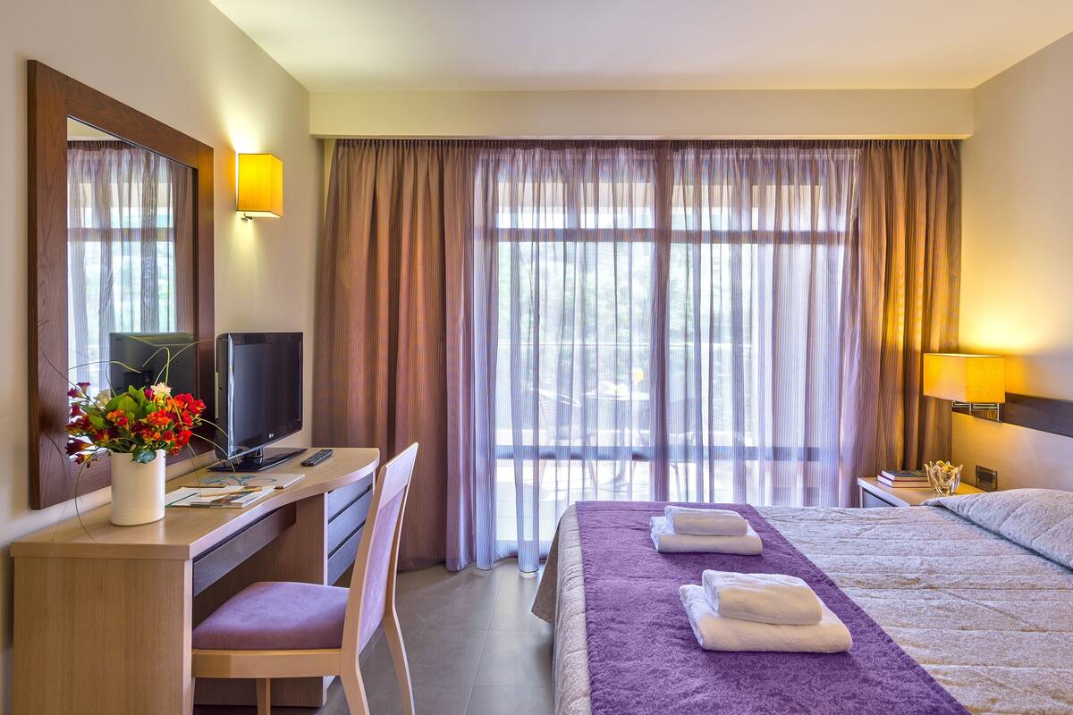 Ein Hotelzimmer mit einem Bett, einem Schreibtisch mit Computer, einem Sessel, einem Fernseher und einem Fenster mit Vorhängen.