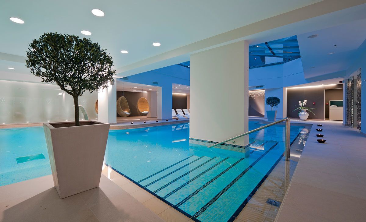 A modern indoor swimming pool with clear lanes, a central tree planter, and contemporary lounge areas.
