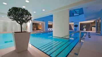 A modern indoor swimming pool with clear lanes, a central tree planter, and contemporary lounge areas.