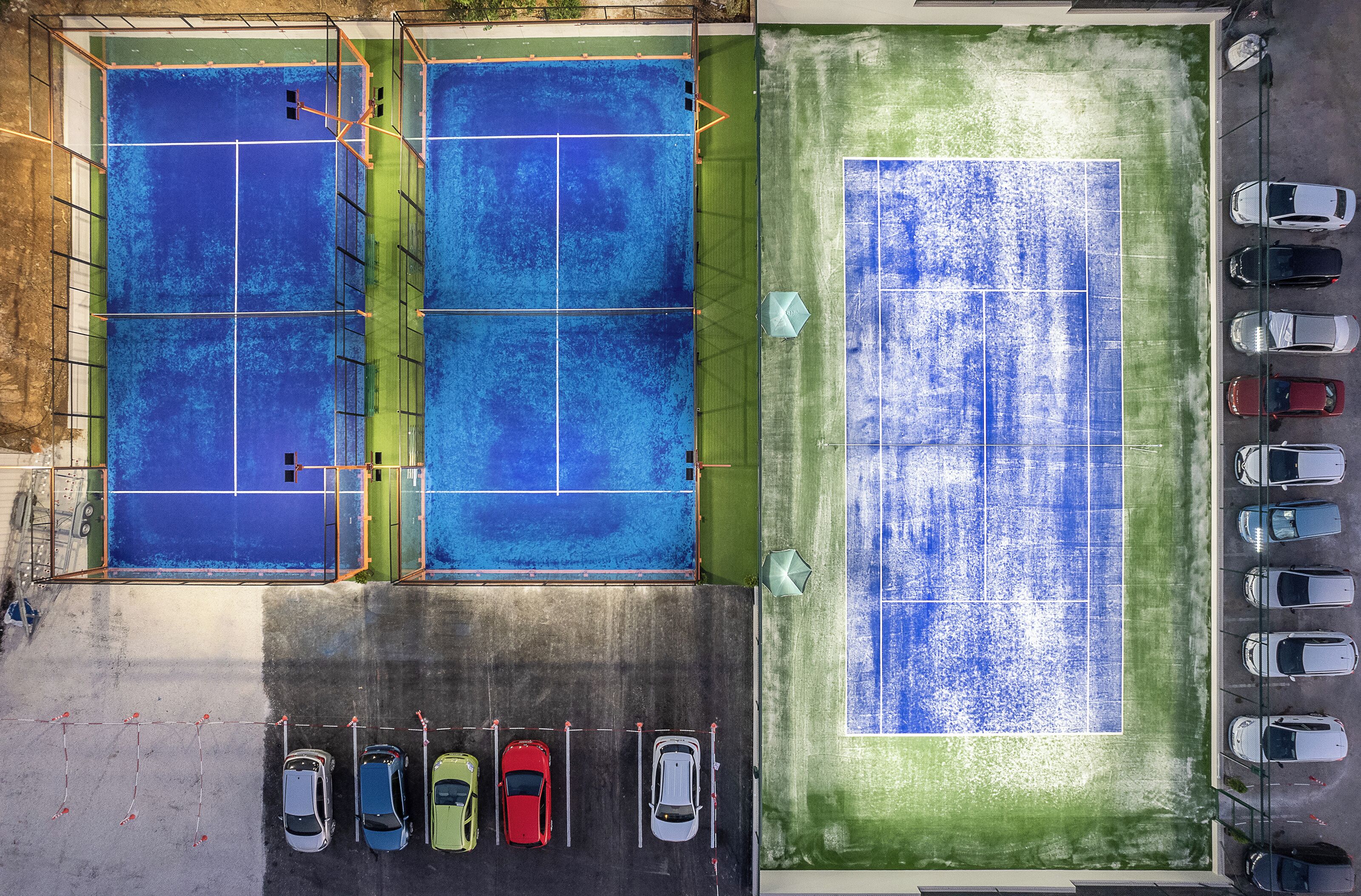 A multi-sport court with tennis and basketball courts, surrounded by a parking area with several cars.