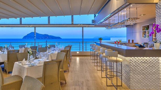 A restaurant with tables set for dining, a bar area, and a view of the ocean. - Porto Platanias Beach Resort & Spa (Platanias)