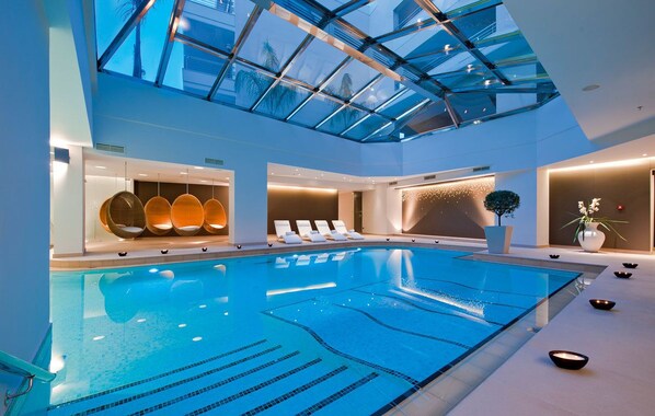 A modern indoor swimming pool with a glass ceiling, lounge chairs, and a spa area.