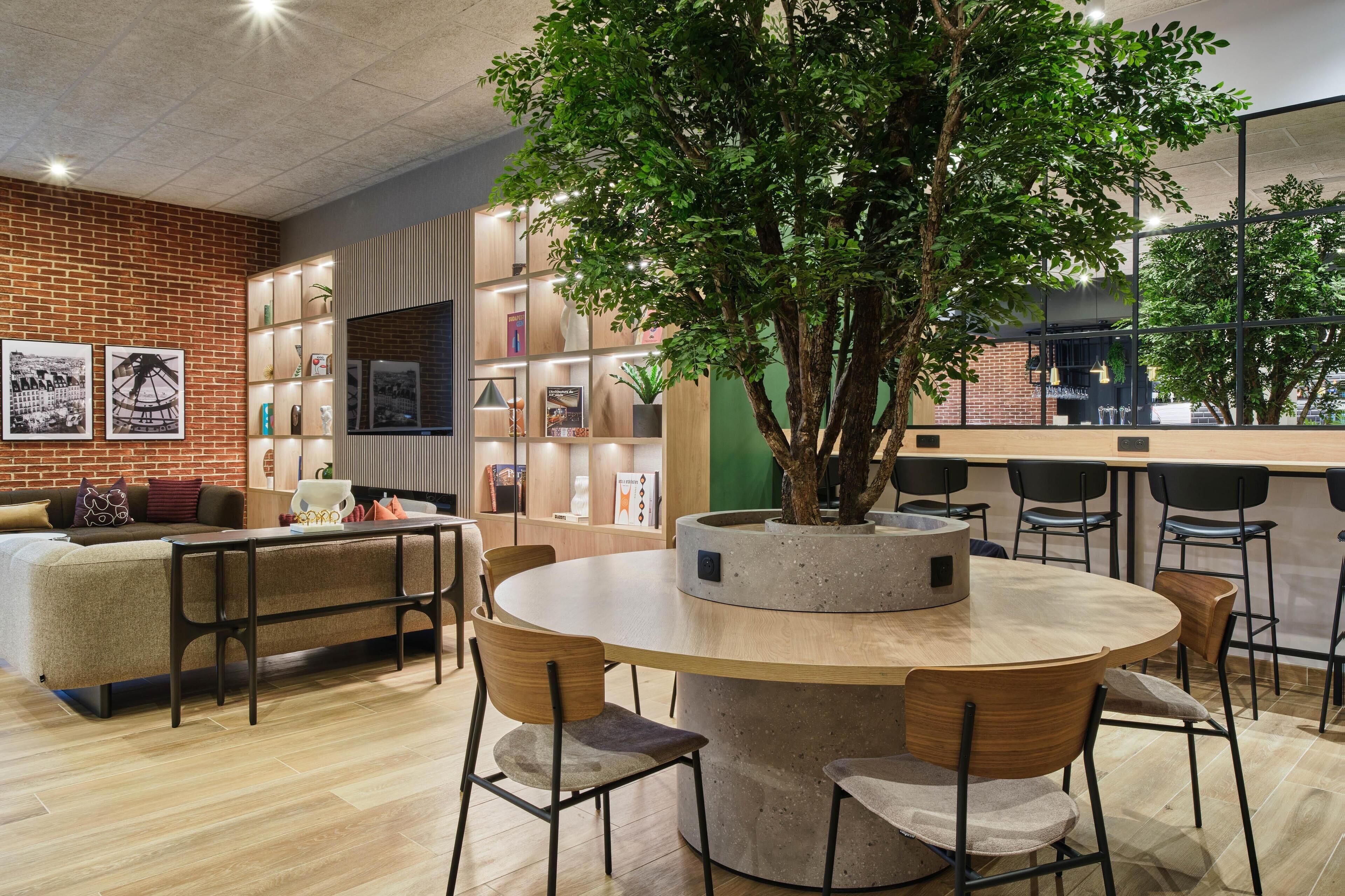 A modern interior with a round wooden table, chairs, a brick wall, and a large tree planter.