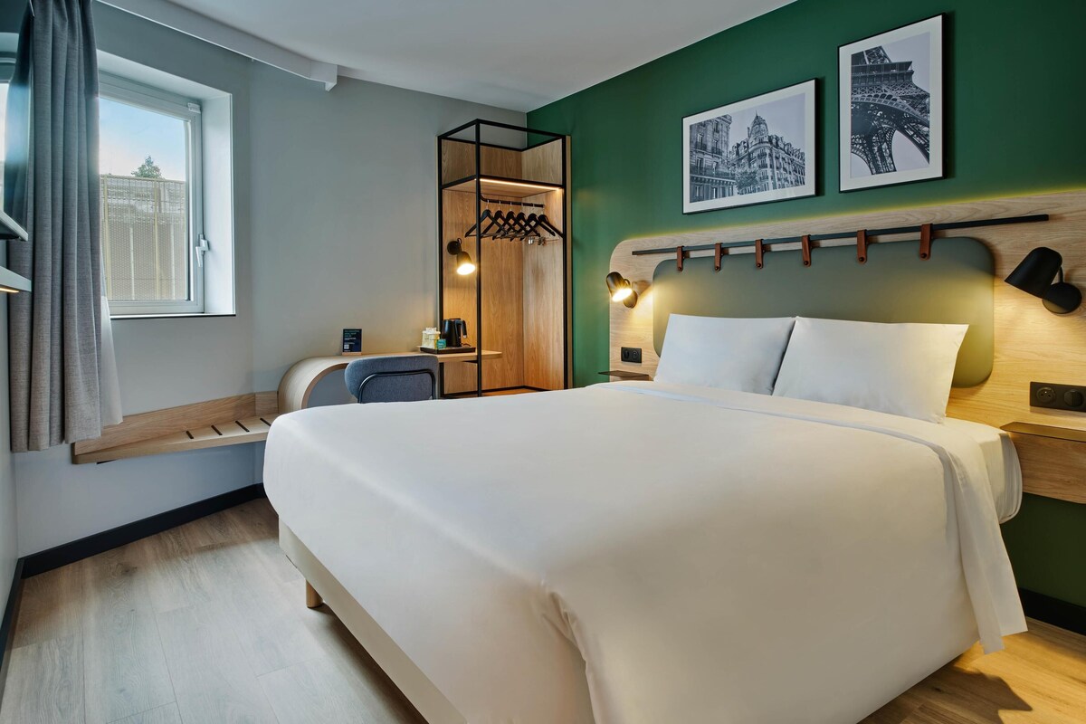 Une chambre d’hôtel avec un lit, un bureau, une chaise, un porte-manteau et deux tableaux encadrés au mur.