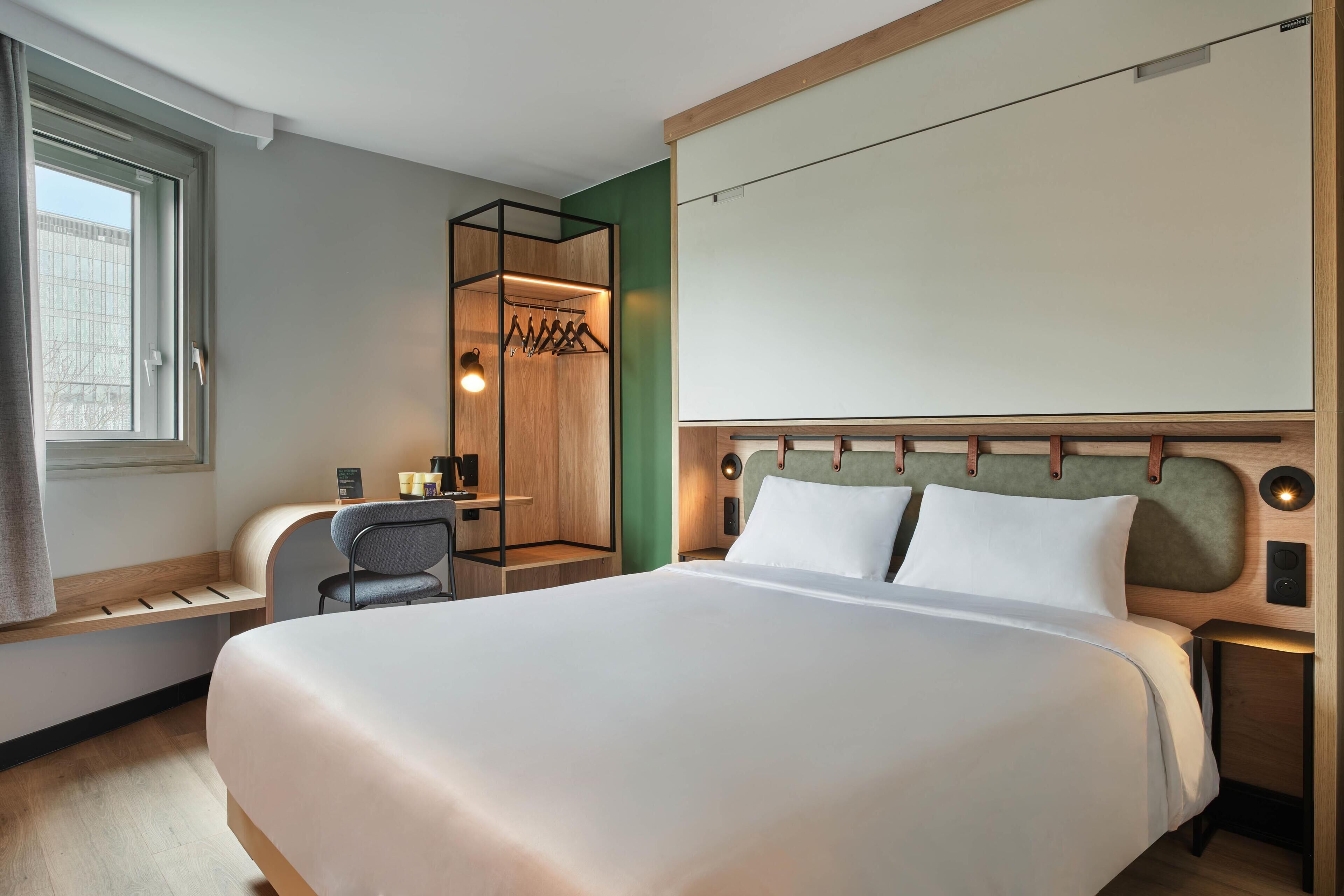 Habitación de hotel con una cama grande, un escritorio con silla, perchero y ventana con cortinas.