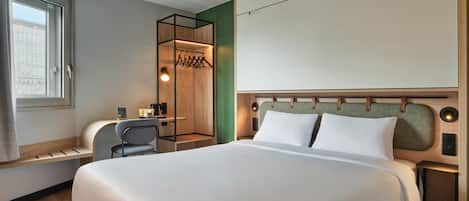 Habitación de hotel con una cama grande, un escritorio con silla, perchero y ventana con cortinas.