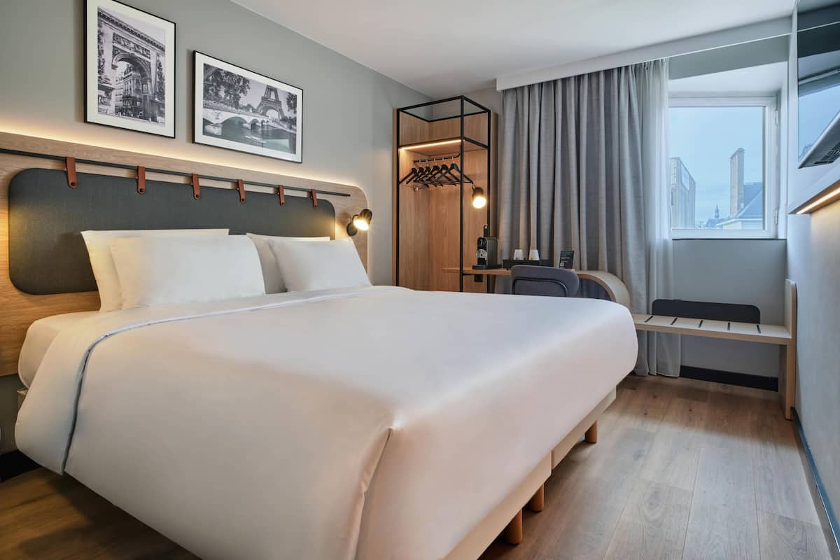 Une chambre d’hôtel avec un grand lit, un bureau avec une chaise, un porte-manteau et une fenêtre avec des rideaux.