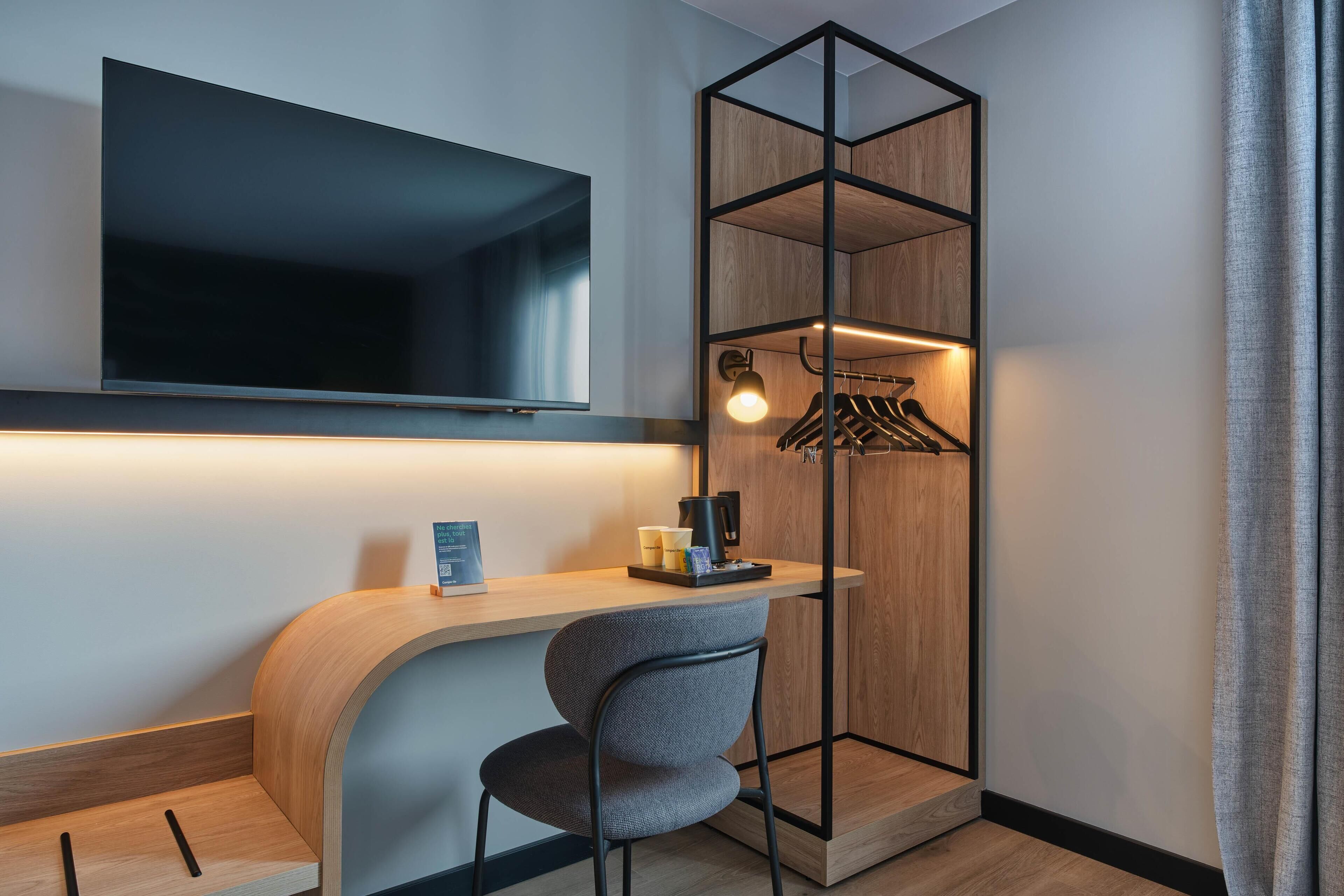 A modern hotel room with a flat-screen TV mounted on the wall, a wooden desk with a chair, a coat rack with hanging clothes, and a small table with a coffee maker and cups.