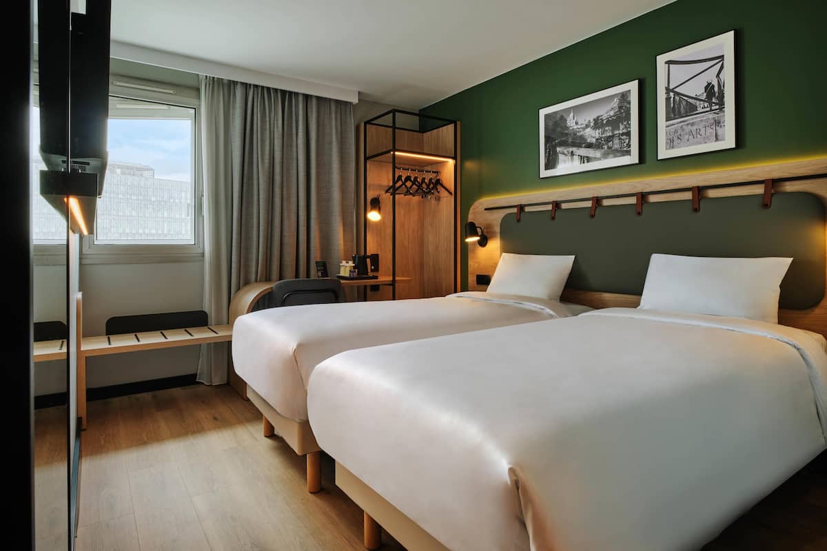 Une chambre d’hôtel avec deux lits, un bureau, une chaise, un porte-manteau et une fenêtre avec des rideaux.