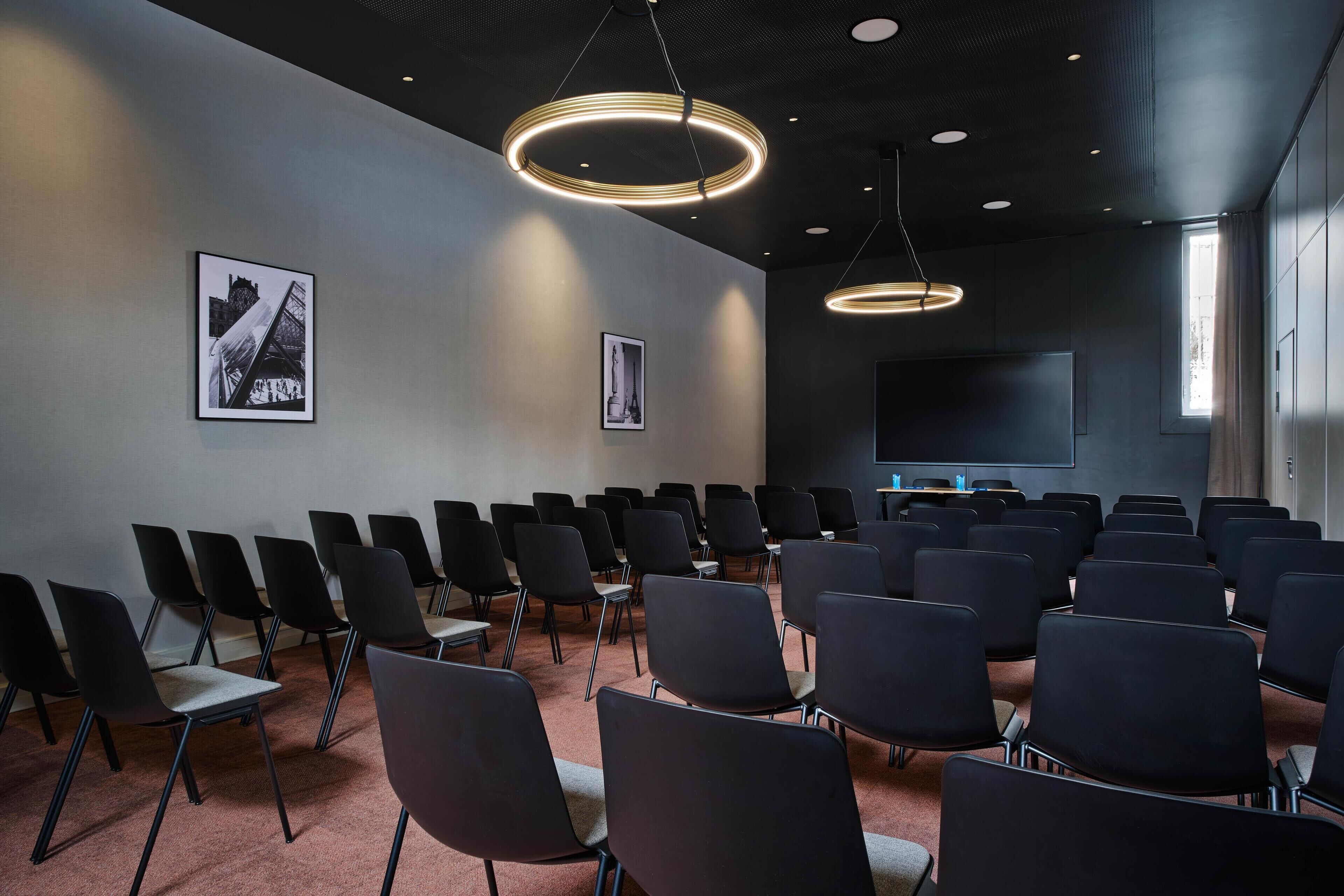 Ein Konferenzraum mit Reihen von Stühlen, einem großen Bildschirm und zwei gerahmten Bildern an der Wand.