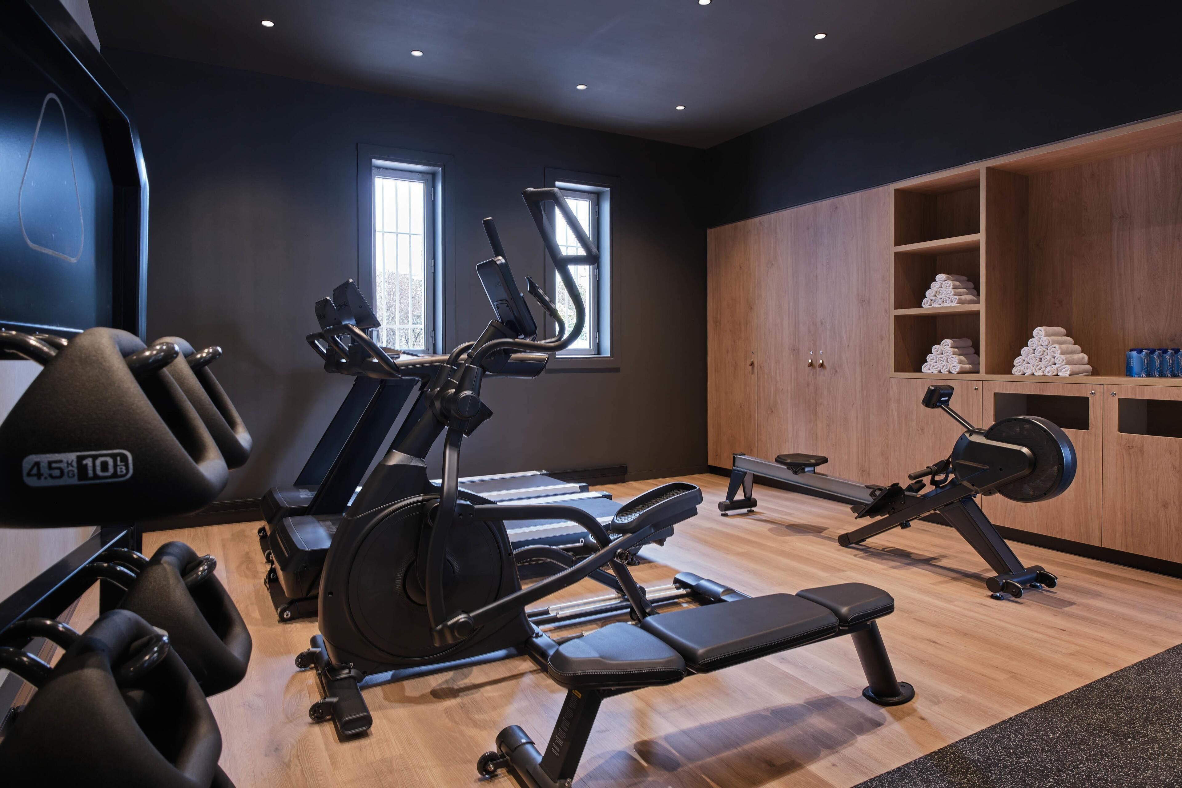 A gym with exercise equipment, a bench, and a wooden cabinet with towels.
