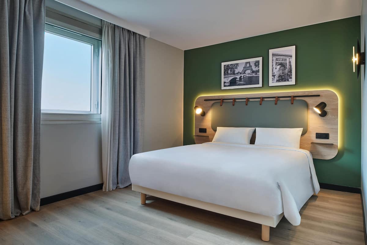 Un lit bien fait dans une pièce dotée d’une grande fenêtre, d’une tête de lit avec des lampes fixées au mur, et de deux tableaux encadrés accrochés au mur.