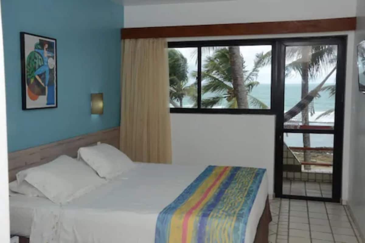 Un dormitorio con cama, ventana con vista a palmeras y al mar, un cuadro en la pared y una cortina.