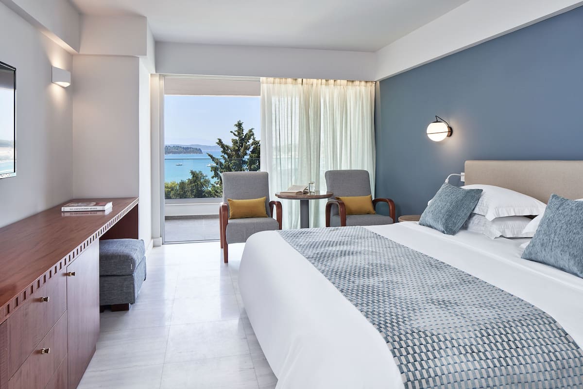 A hotel room with a large bed, a desk, two chairs, a small table, and a view of the sea.