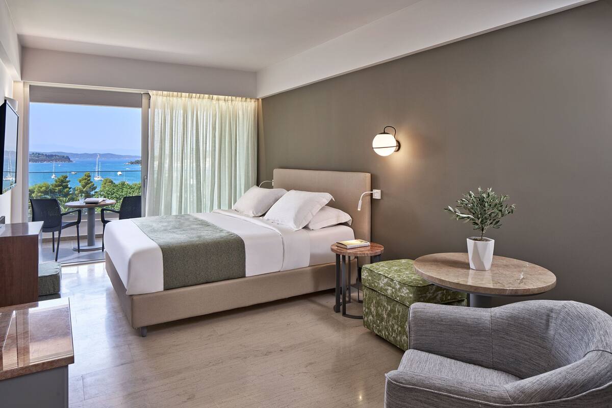 A modern hotel room with a bed, a desk, a chair, a small table with a plant, and a view of the sea.