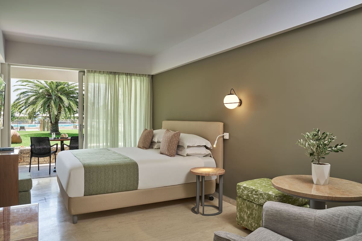 A modern hotel room with a bed, a round table, a potted plant, and a view of palm trees through the window.