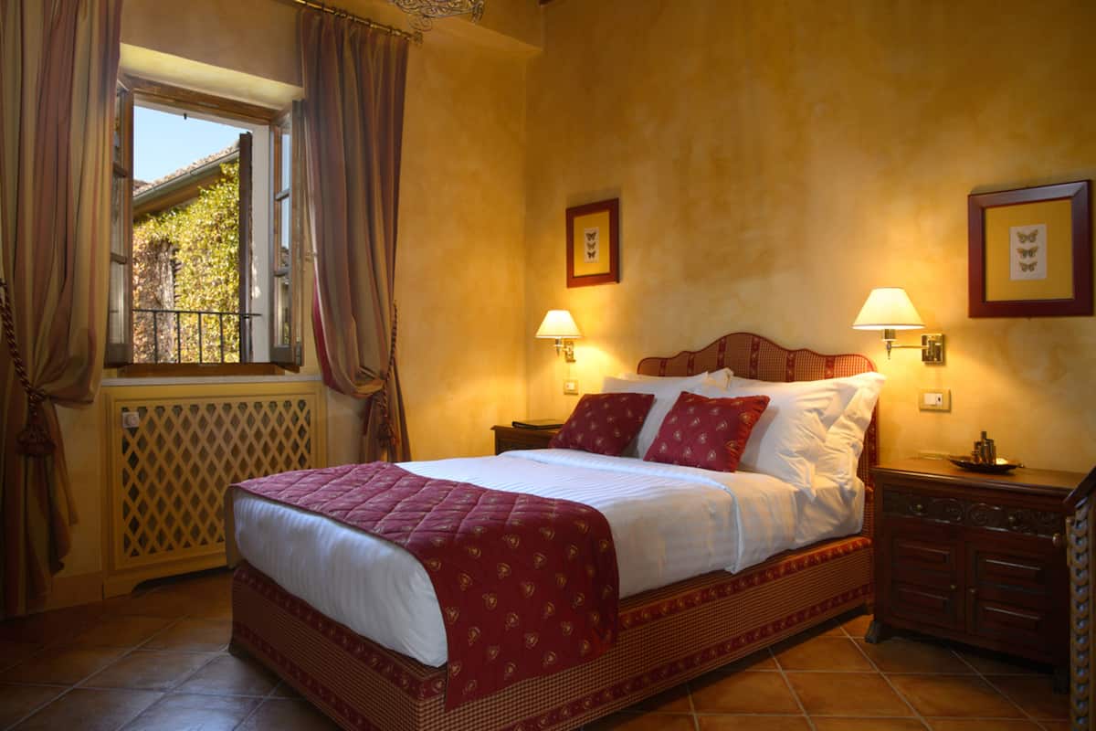 A bedroom with a large bed, two bedside lamps, a window with curtains, and a radiator.