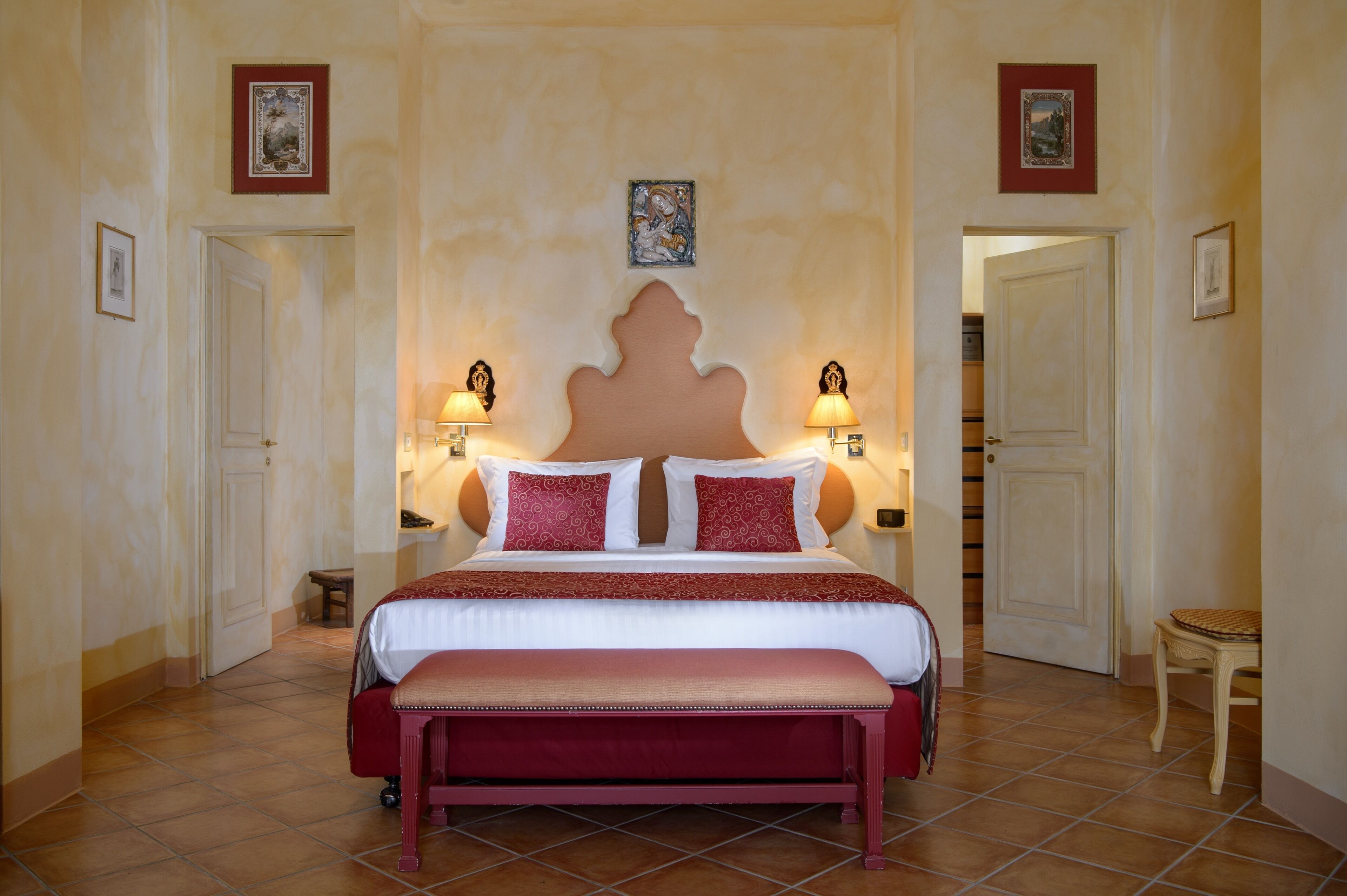 A bedroom with a large bed, two bedside lamps, a bench, and a door leading to another room.