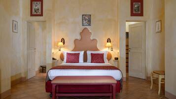 A bedroom with a large bed, two bedside lamps, a bench, and a door leading to another room.