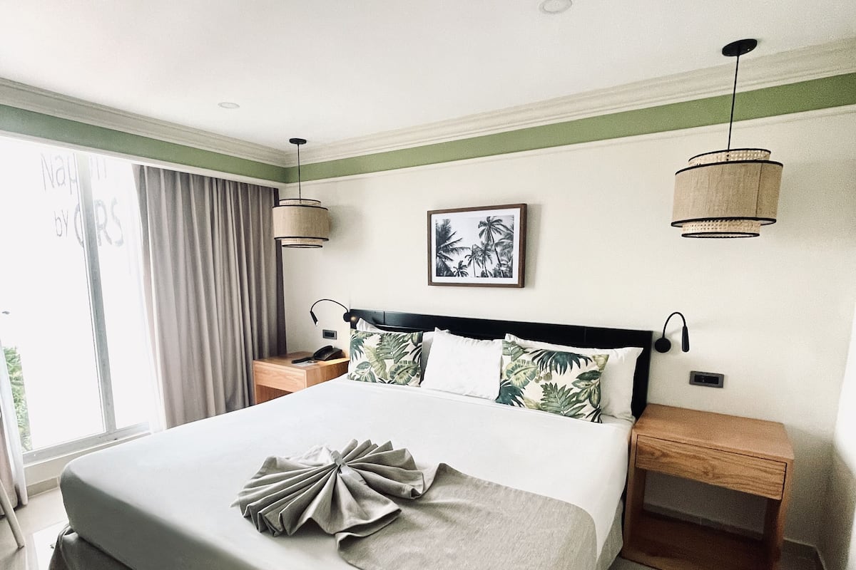 Une chambre d’hôtel bien rangée, avec un grand lit, une table de chevet, une petite table d’appoint, une lampe fixée au mur et un tableau encadré accroché au mur.