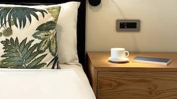 Una cama impecable con un cojín estampado con hojas tropicales, una mesita de noche con una taza blanca y un libro, y una lámpara fijada a la pared.
