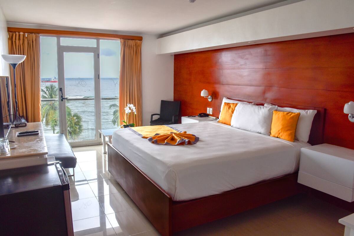Une chambre d’hôtel avec un grand lit, un bureau avec une lampe, une chaise et une vue sur la mer par la fenêtre.