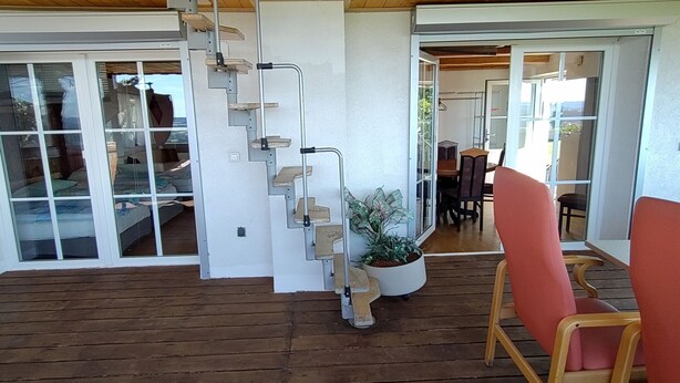 A wooden deck with a staircase leading to a glass door, a potted plant, and a dining area with chairs. - Geinberg Suites & Vianova Lodges (Polling im Innkreis)
