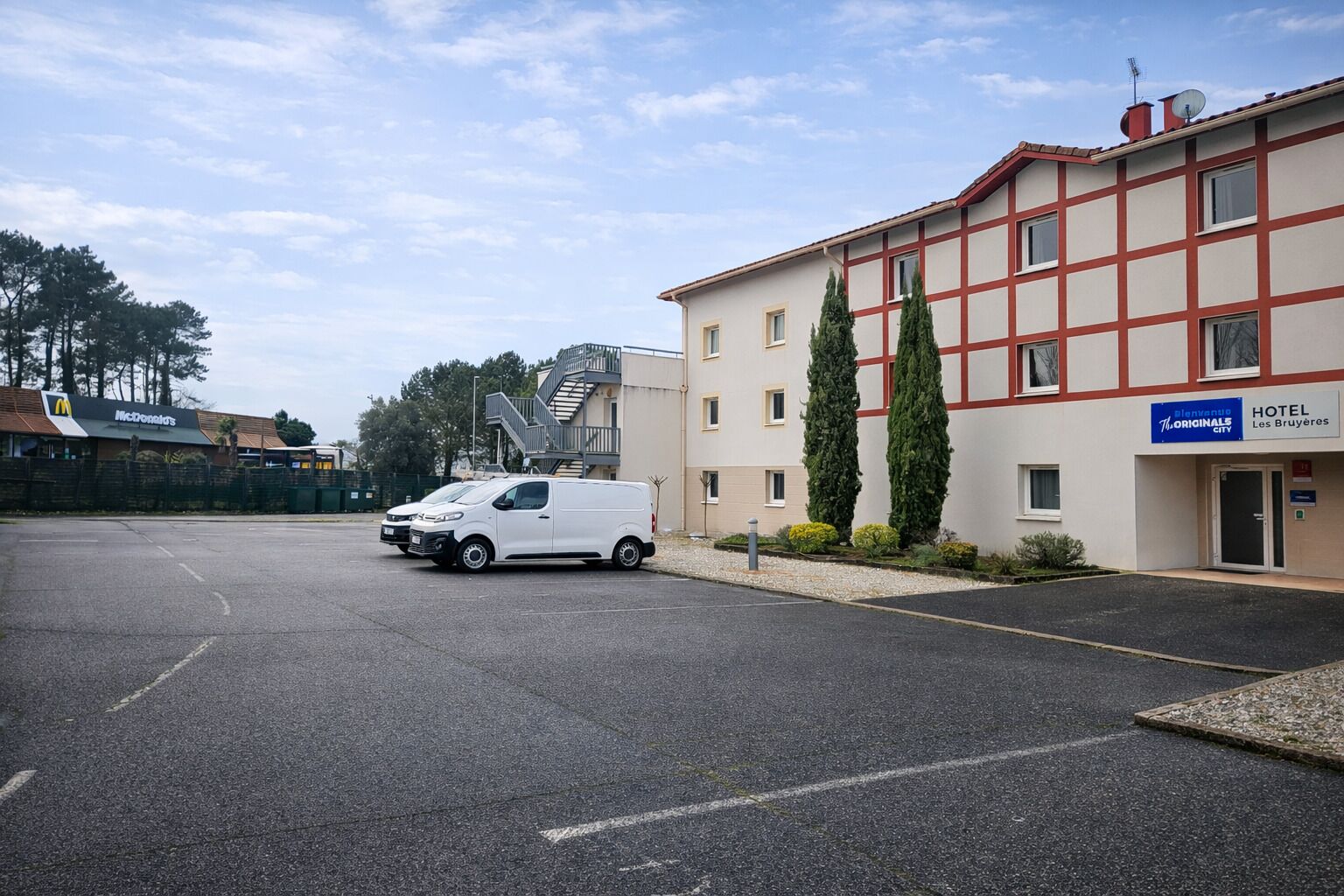 A white van parked in a parking lot, a hotel building with a sign reading "Hotel Les Bruyères," and a McDonald's restaurant in the background.