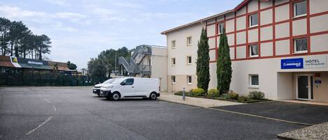 Un fourgon blanc est garé sur un parking, un bâtiment d’hôtel avec une enseigne indiquant « Hôtel Les Bruyères », et un restaurant McDonald’s en arrière-plan.