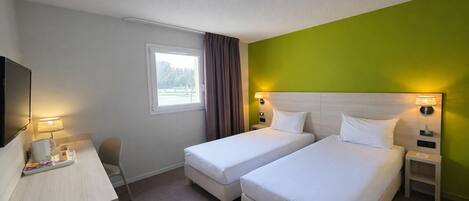Ein Hotelzimmer mit zwei Betten, einem Schreibtisch mit Lampe, einem Fernseher und einem Fenster mit Vorhängen.