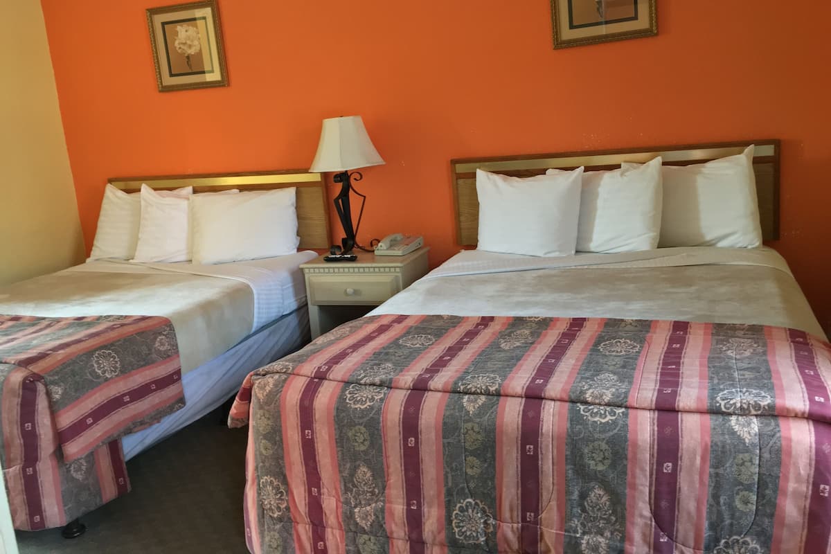 A hotel room with two beds, each with white linens and a patterned bedspread. There are two bedside tables with lamps and a phone. The walls are orange, and there are two framed pictures on the wall.