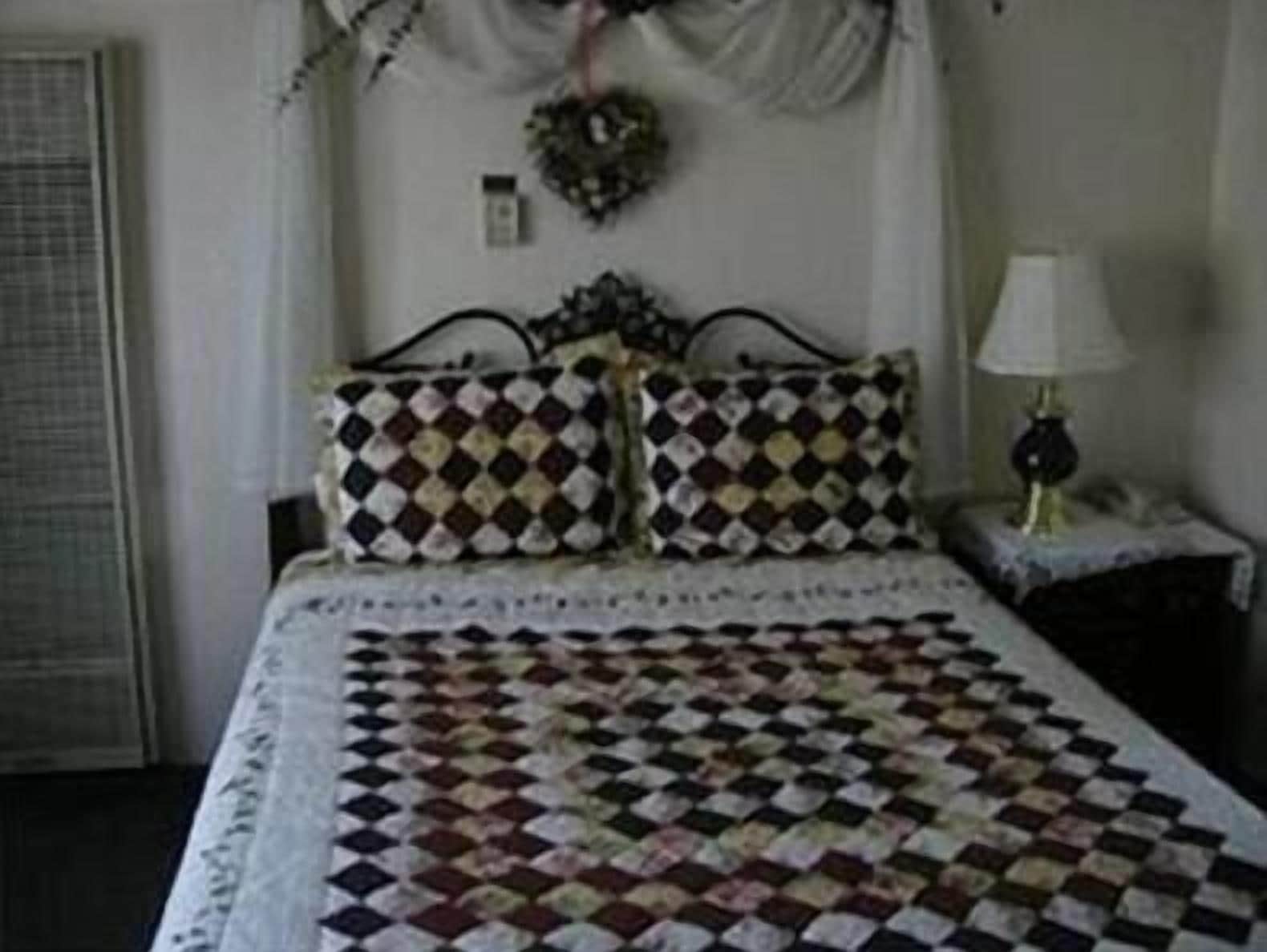 A bedroom with a quilted bedspread, two patterned pillows, a nightstand with a lamp, and a wall-mounted light switch.