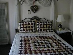 Quarto com colcha acolchoada, duas almofadas estampadas, mesa de cabeceira com abajur e interruptor de luz fixado na parede.