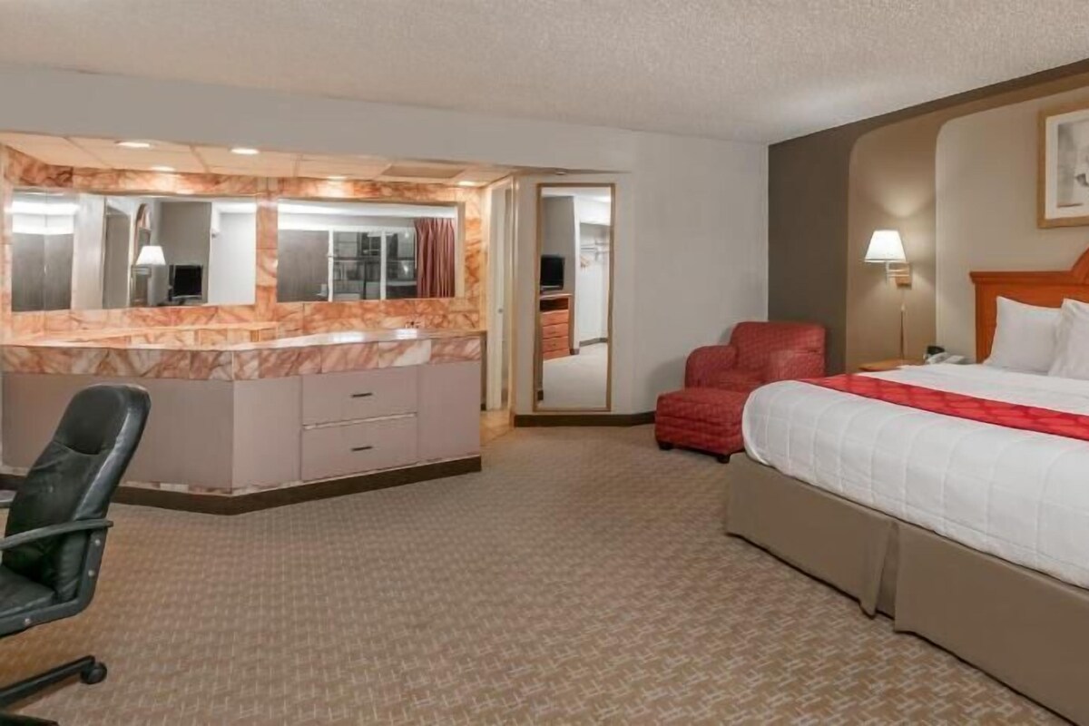 A hotel room with a bed, a desk with a mirror, a chair, and a red armchair.